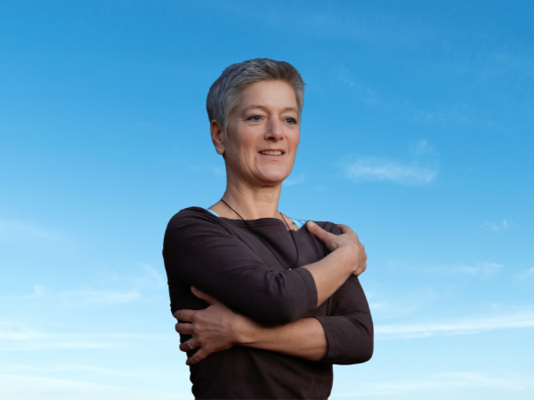 Vera Wollenweber vor blauem Himmel, die Arme liebevoll um sich selbst gelegt – sich selbst halten als Beginn von Empfangen lernen