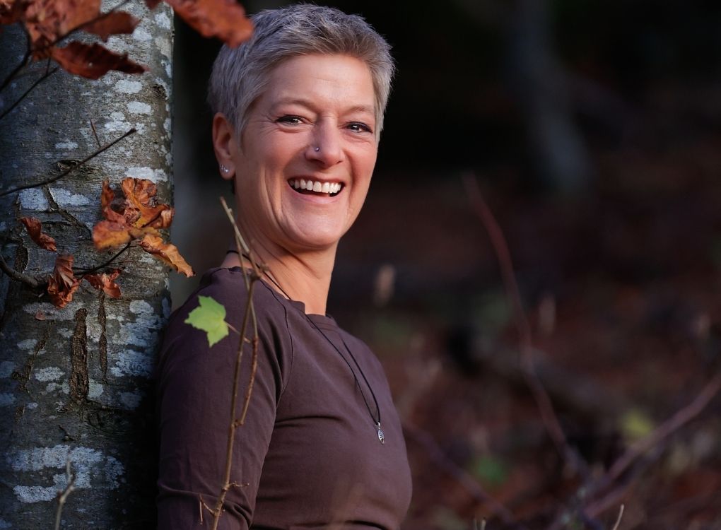 Vera lehnt an einem Baum im Herbstwald und lacht strahlend in die Kamera.