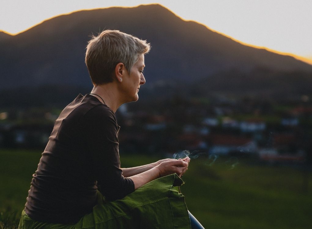 Vera sitzt im Sonnenuntergang, ein Berg ist im Hintergrund von der Sonne beleuchtet. "Ich bin verbunden mit den geistigen Kräften und die Energien fließen."