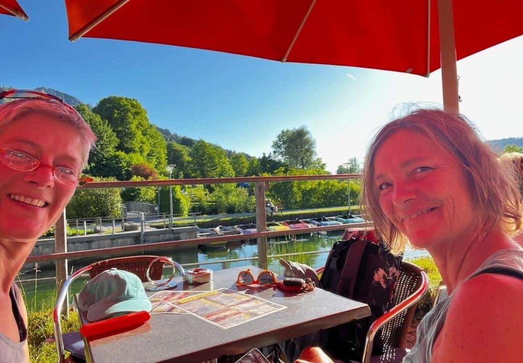 Conny und Vera sitzen lächelnd im Cafe am Alpsee, um eine Erfrischung zu bestellen.