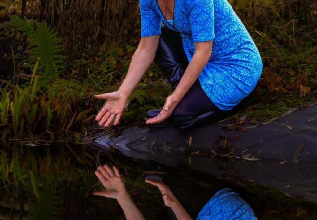 Vera kniet in einem blauen Kleid am Ufer eines Weihers, ihre geöffneten Hände spiegeln sich im dunklen Wasser.